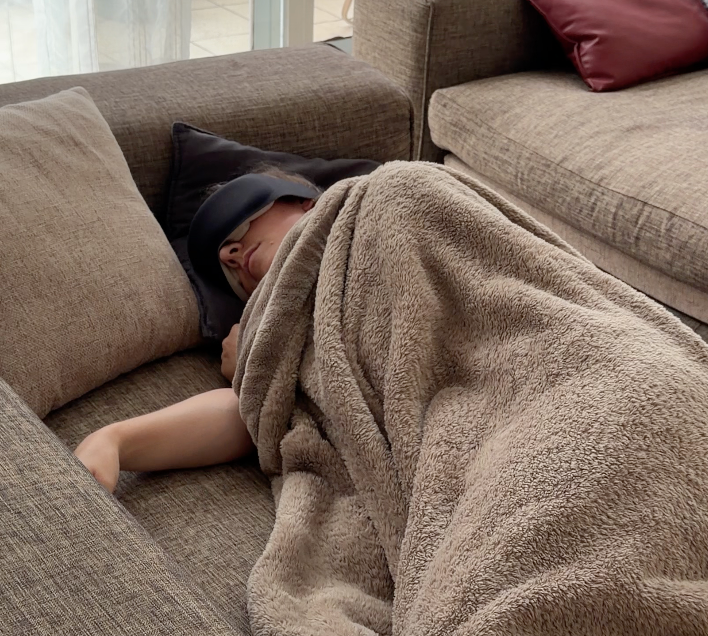 Person asleep on their side on a sofa under a blanket, wearing the Gravity weighted sleep mask